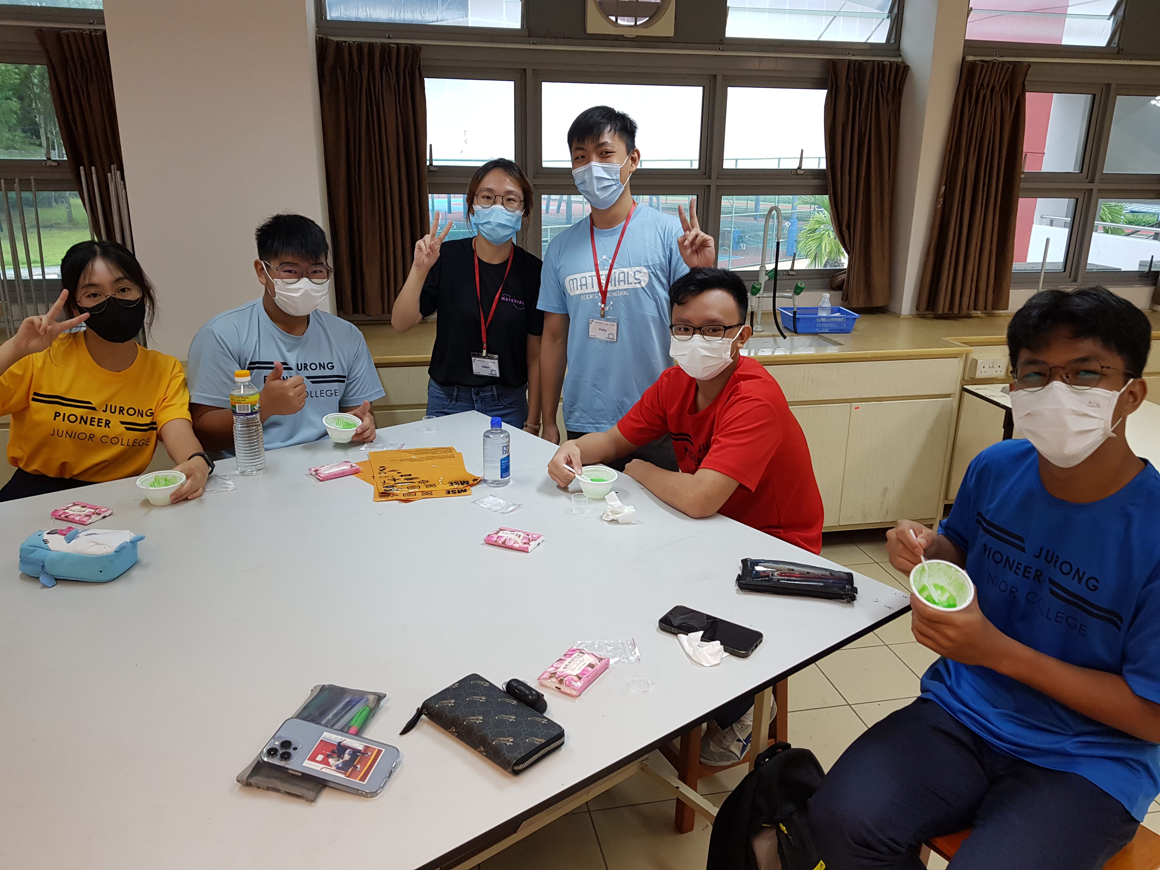 Six masked students around a classroom table, some flashing peace signs, eating green dessert from cups.